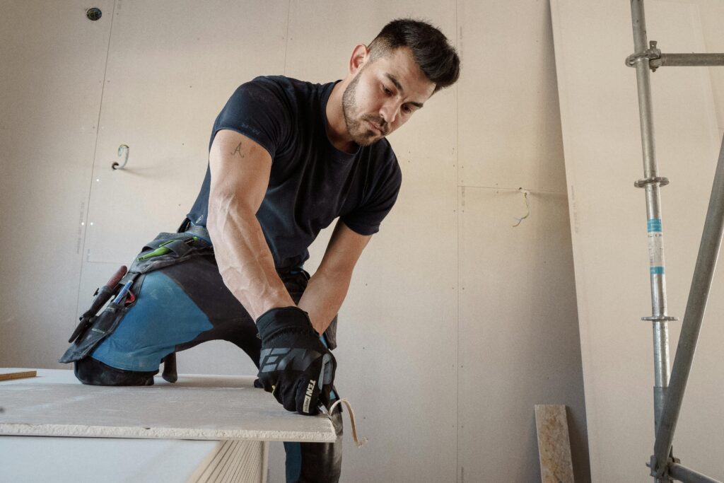 Man in gloves cutting plasterboard for drywall installation inside a building.